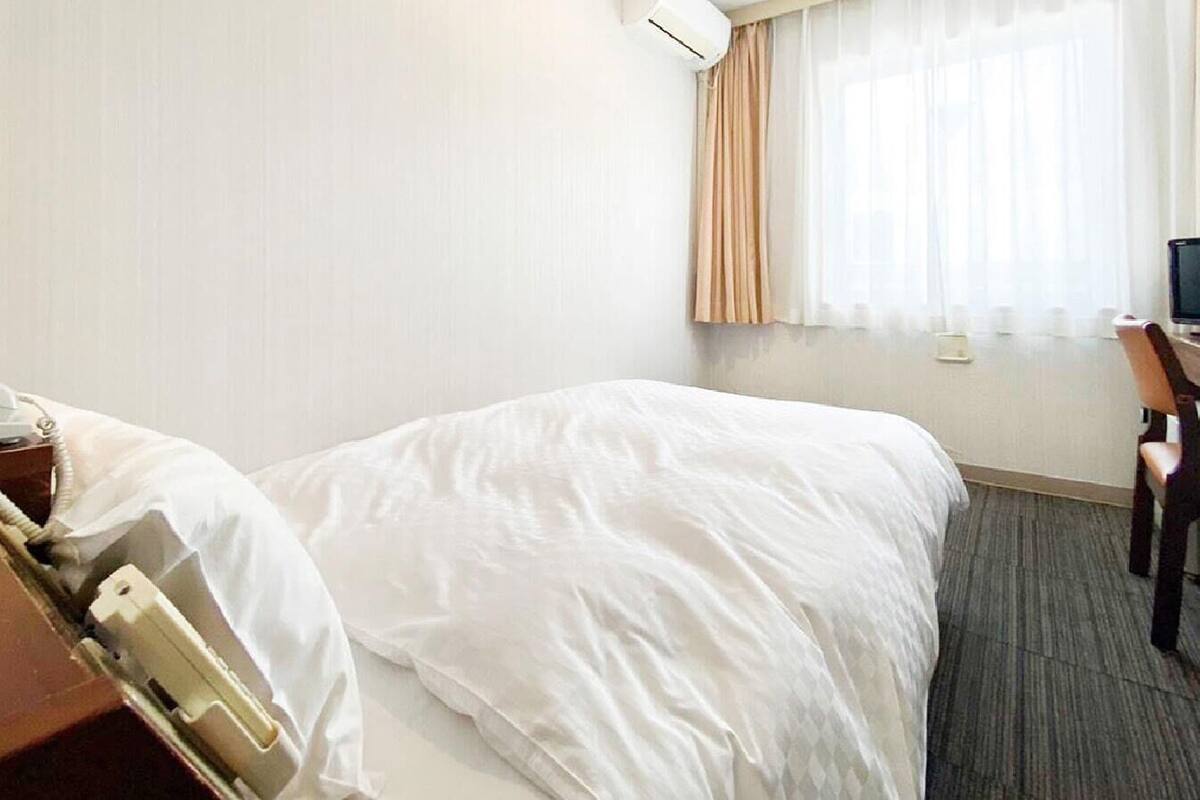 Ein Hotelzimmer mit einem Bett, einem Schreibtisch mit Computer, einem Telefon und einem Fenster mit Vorhängen.