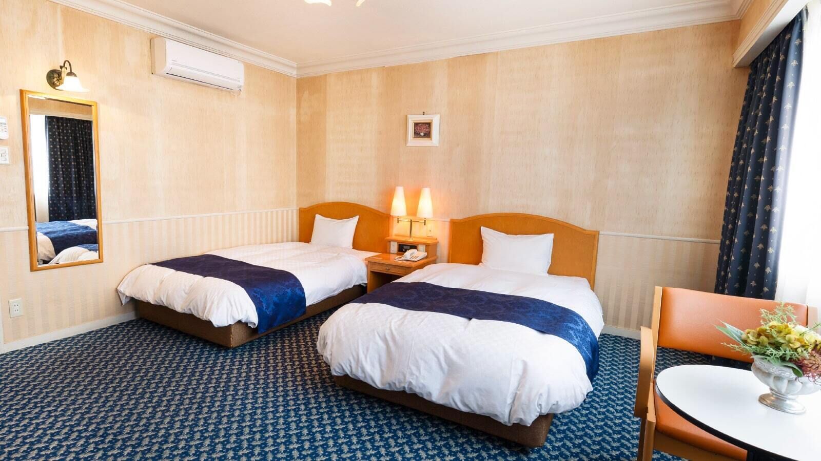 Quarto de hotel com duas camas, uma mesa de cabeceira com abajur, um espelho, uma pequena mesa com um vaso de flores e um tapete estampado.