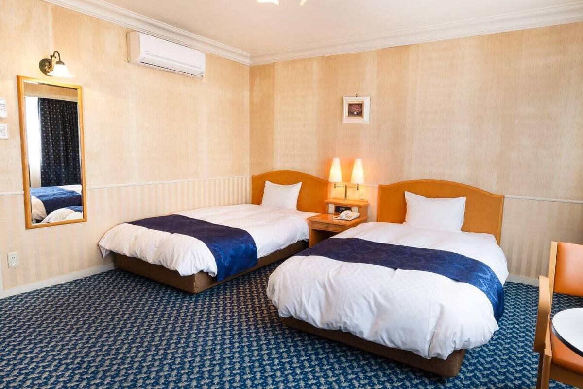 Ein Hotelzimmer mit zwei Betten, einem Nachttisch mit Lampe, einem Spiegel, einem kleinen Tisch mit Blumenvase und einem gemusterten Teppich.