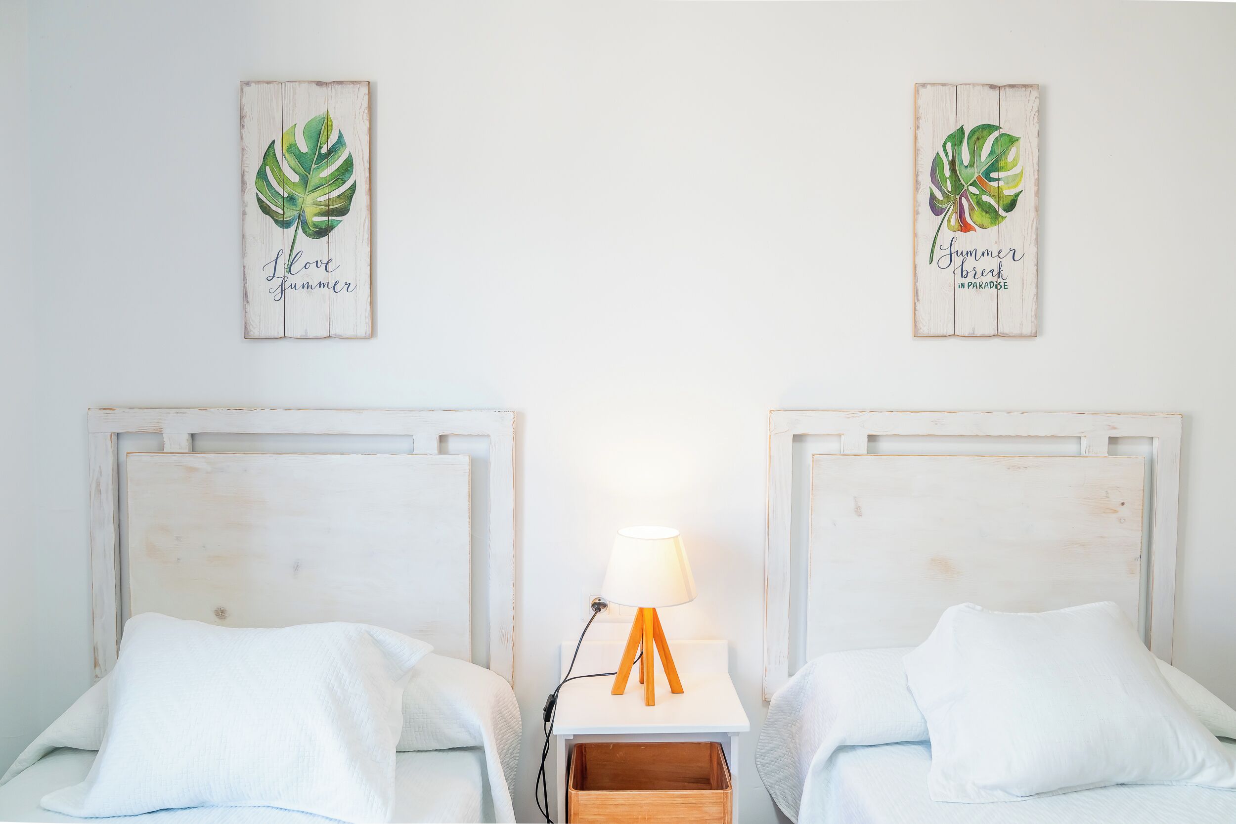 Deux lits avec du linge de lit blanc, une table de chevet avec une lampe et deux œuvres d’art murales représentant des feuilles tropicales.