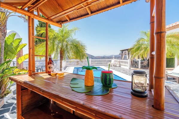 Una barra de madera con vistas a una piscina y montañas, rodeada de palmeras.