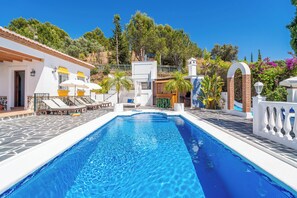Una piscina con sillones de descanso, un edificio blanco con detalles amarillos y montañas al fondo.