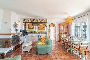 Una sala de estar con chimenea, sofá verde, mesa de comedor con sillas, cocina al fondo y un gran ventanal.