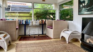 A cozy room with wicker furniture, a bookshelf, a fireplace, and large windows with a view of greenery. - Beautiful quiet 2 persons caravan on the Lauwersmeer (Anjum)