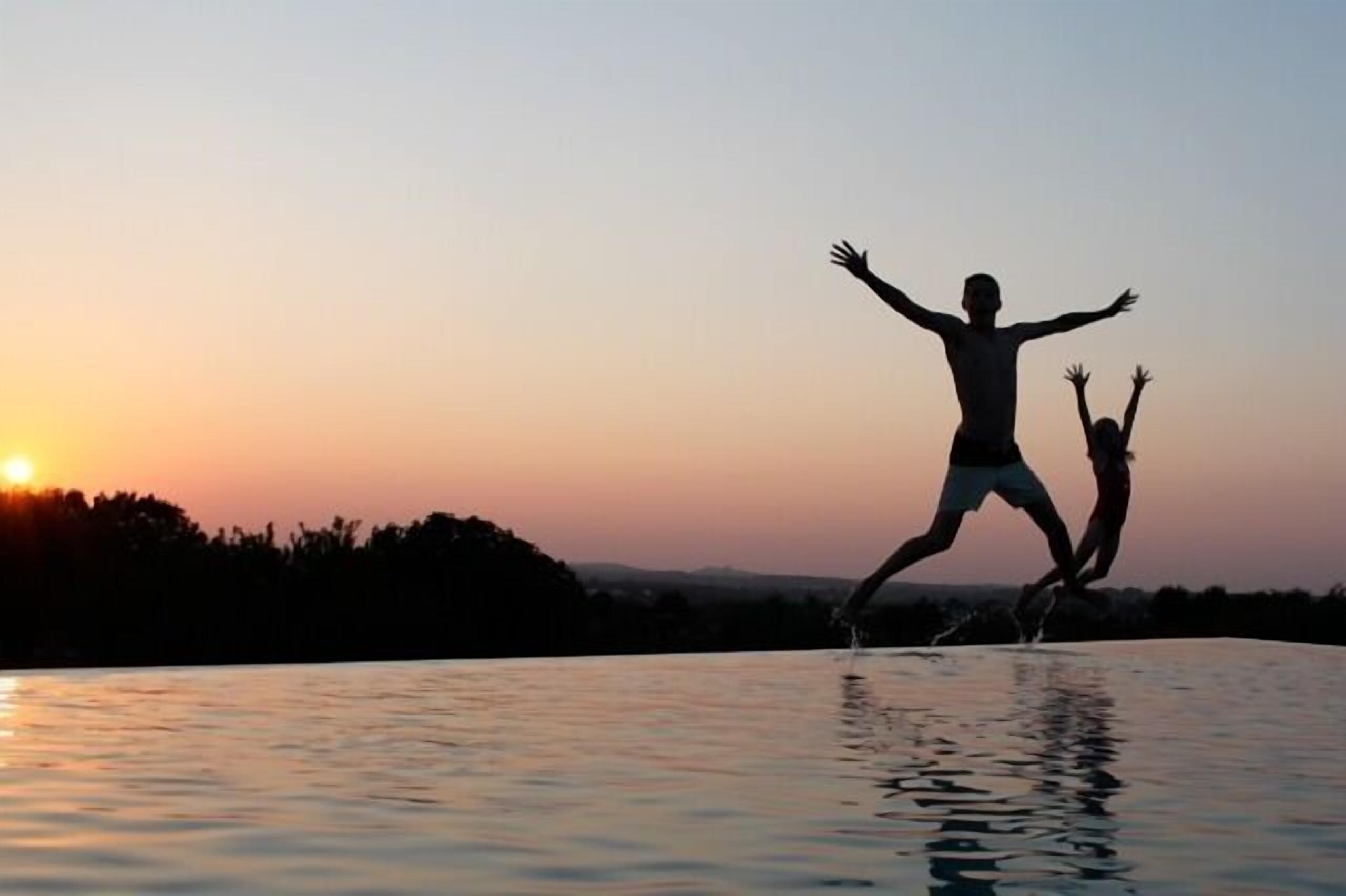 Two people jumping into a pool at sunset with a clear sky and distant trees.
