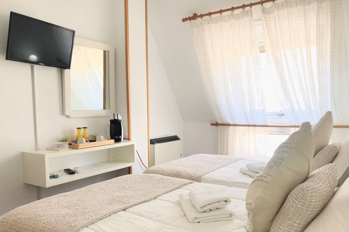 Een slaapkamer met een aan de muur bevestigde televisie, een nachtkastje met een koffiezetapparaat, een bed met witte beddengoed en kussens, en een raam met dunne gordijnen.