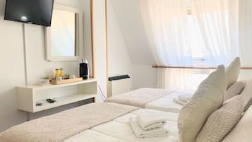 Un dormitorio con una televisión montada en la pared, una mesita de noche con cafetera, una cama con ropa de cama y almohadas blancas, y una ventana con cortinas traslúcidas.