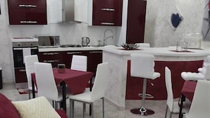 Una cocina moderna con armarios blancos y encimera roja. Hay un comedor con sillas blancas y sofá rojo.