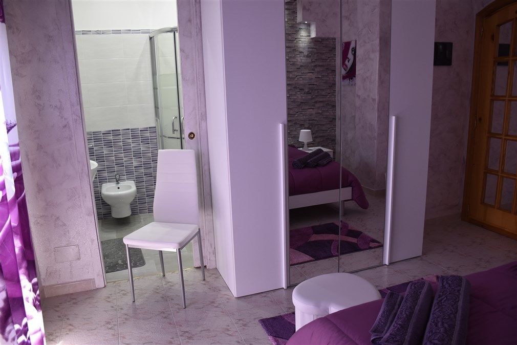 A bedroom with a bed, a chair, and a small table. There is a bathroom visible through a glass door.