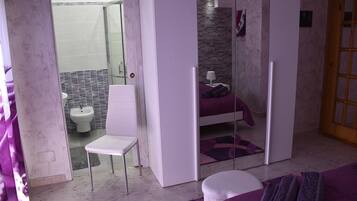 A bedroom with a bed, a chair, and a small table. There is a bathroom visible through a glass door.