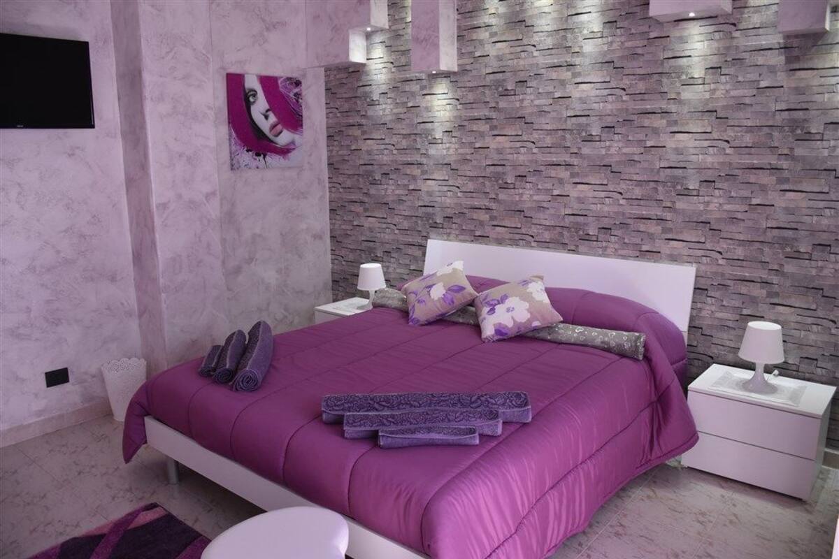 Ein modernes Schlafzimmer mit Wand im Steinoptik, einer violetten Bettdecke und einem Nachttisch mit Lampe.