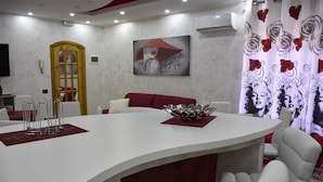 Una sala de estar moderna con una mesa de centro blanca curvada, cojines rojos y un cuenco de cristal decorativo. En la pared hay un cuadro de una mujer con un sombrero rojo, y se puede ver una cortina con estampado de rosas.