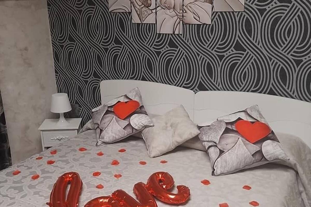 Ein Bett mit einem gemusterten Kopfteil, Kissen mit Herzverzierungen und roten, herzförmigen Ballons, die das Wort „love“ bilden.