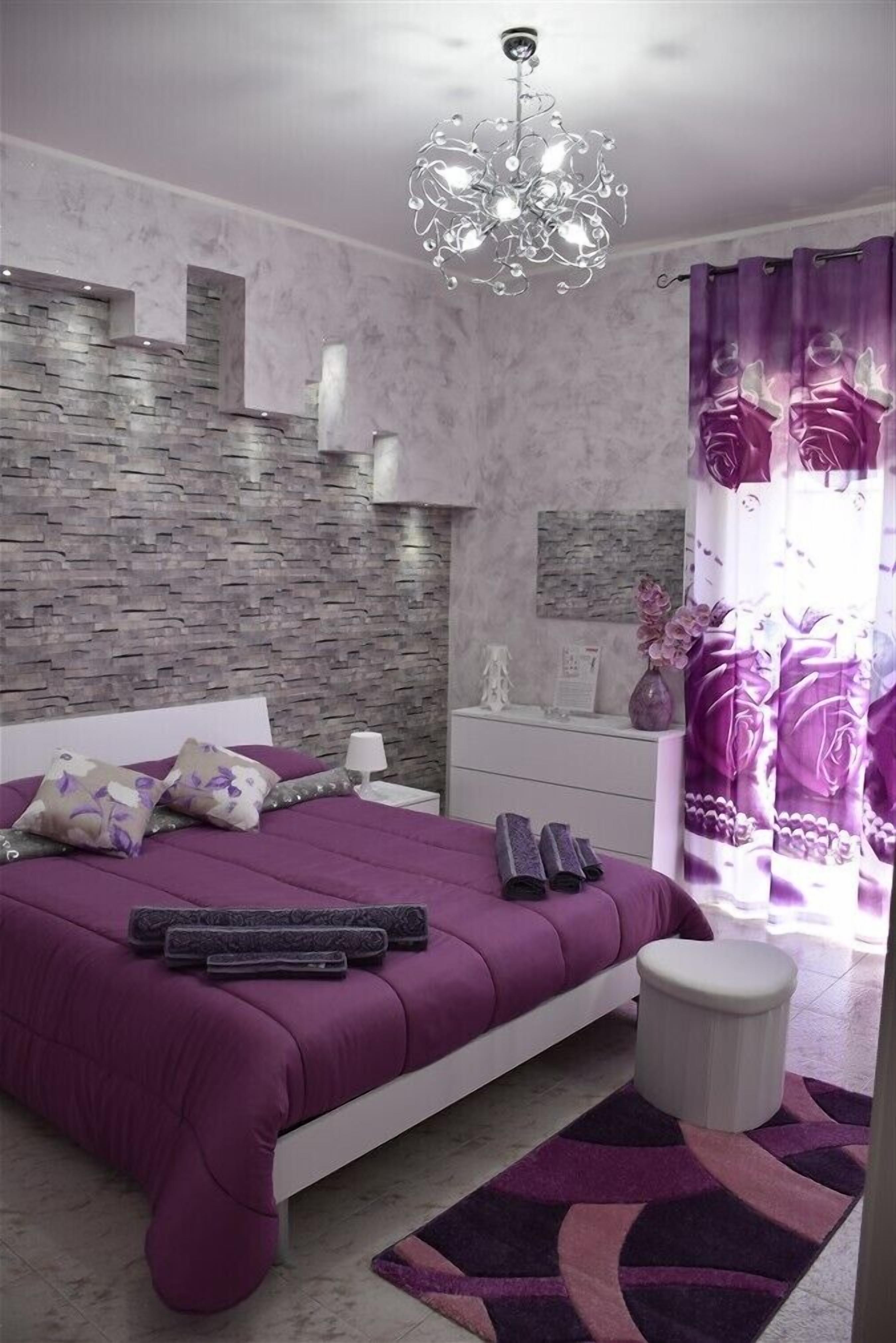A modern bedroom with a stone wall, a purple bedspread, and a chandelier.