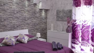 A modern bedroom with a stone-patterned headboard, a large bed with purple bedding, a white nightstand, a white bedside table, and a decorative chandelier.
