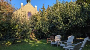 A garden with a brick building, green bushes, and Adirondack chairs.