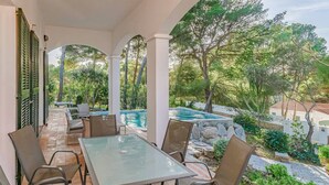 Un patio al aire libre con una mesa y sillas de vidrio, una piscina y un jardín con árboles y arbustos.