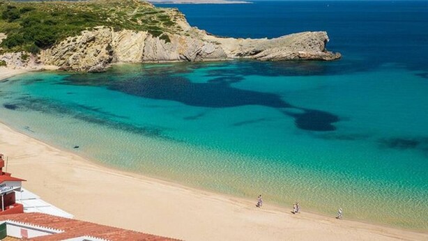 Una playa costera con aguas turquesas cristalinas, un acantilado rocoso y algunas personas caminando sobre la arena.