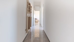 A narrow hallway with a hexagonal patterned floor, white walls, and a door at the end leading to a room with a bed and a window.