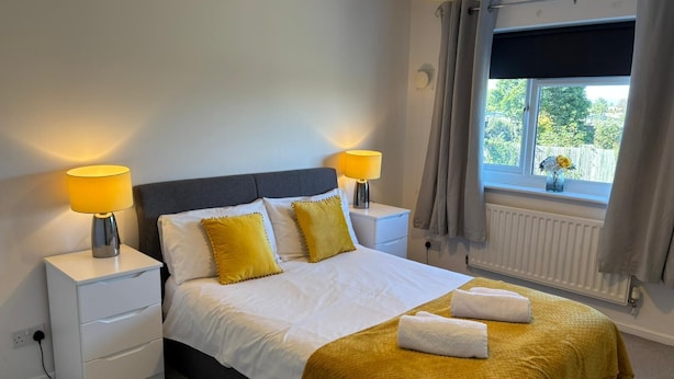A neatly made bed with white and yellow bedding, two bedside lamps, a window with curtains, and a radiator. - HEREFORD – Kestrel House: 4-Bed Home North of City, Parking for 2, Sleeps 7 (Hereford)