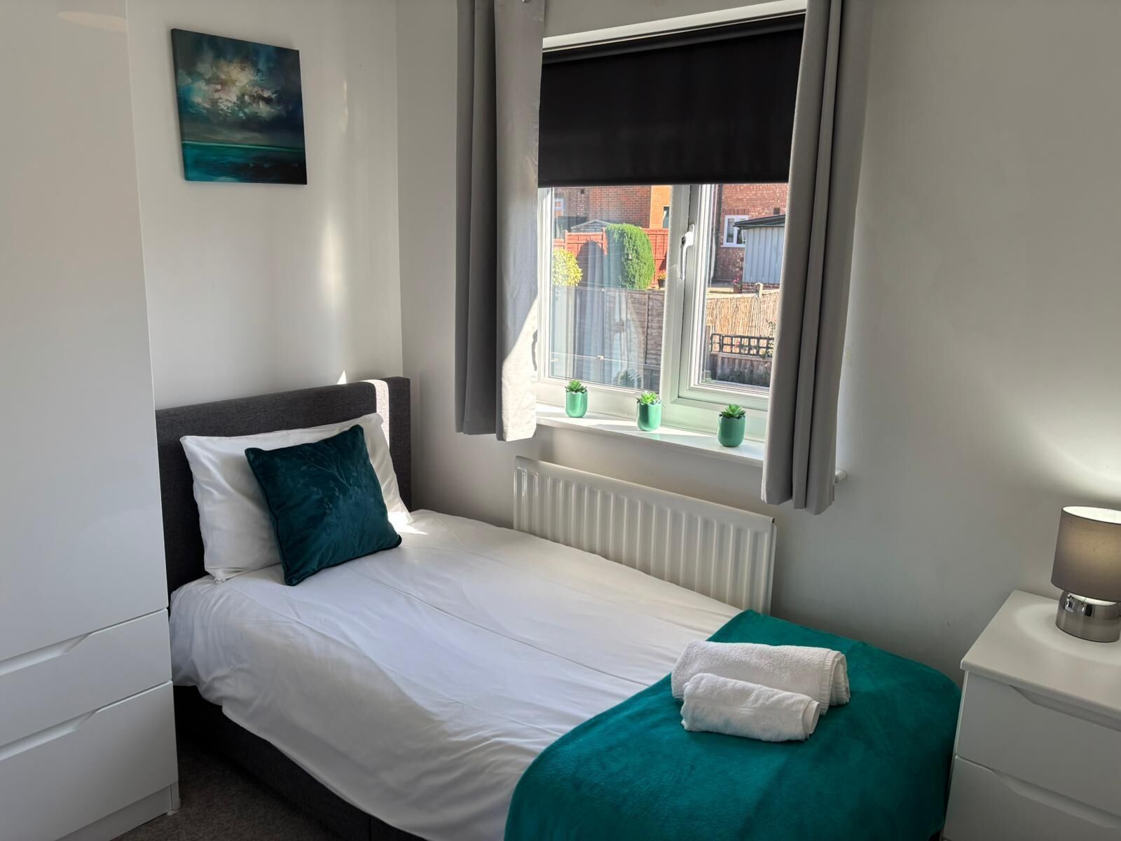 Cama bem arrumada com roupa de cama branca e azul-turquesa, um cobertor azul-turquesa e um travesseiro branco. Tem uma mesinha de cabeceira com abajur, e uma janela com vista para um jardim e edifícios.