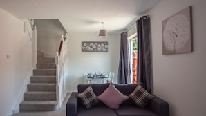 A living room with a dark sofa, a dining table with chairs, and a staircase leading to another level. - HEREFORD – Kestrel House: 4-Bed Home North of City, Parking for 2, Sleeps 7 (Hereford)