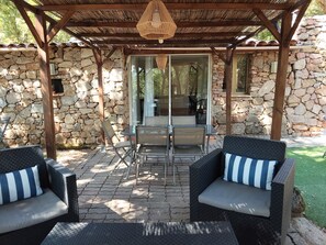 A covered patio with stone walls, a glass door leading inside, and outdoor furniture. - COMFORTABLE CHALET HEATED POOL BEACHES AND RIVER 10 MIN AWAY (Conca)