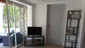A living room with a TV on a wooden stand, a bookshelf, and a sliding glass door leading to an outdoor seating area. - COMFORTABLE CHALET HEATED POOL BEACHES AND RIVER 10 MIN AWAY (Conca)