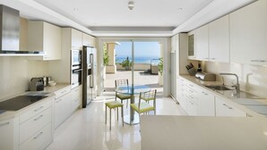 A modern kitchen with a central island, a dining area with chairs, and a view of the ocean through large sliding doors. - 405 Luxury 4 Bed Penthouse, Heated 20m Pool-Front Line-Paseo-Bikes-BBQ-Hot Tub (Estepona)