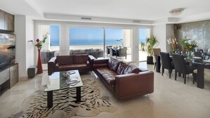 A spacious living room with a large brown leather sofa, a glass coffee table, and a zebra-patterned rug. There is a dining area with a table and chairs, and a large window offering a view of the ocean. - 405 Luxury 4 Bed Penthouse, Heated 20m Pool-Front Line-Paseo-Bikes-BBQ-Hot Tub (Estepona)