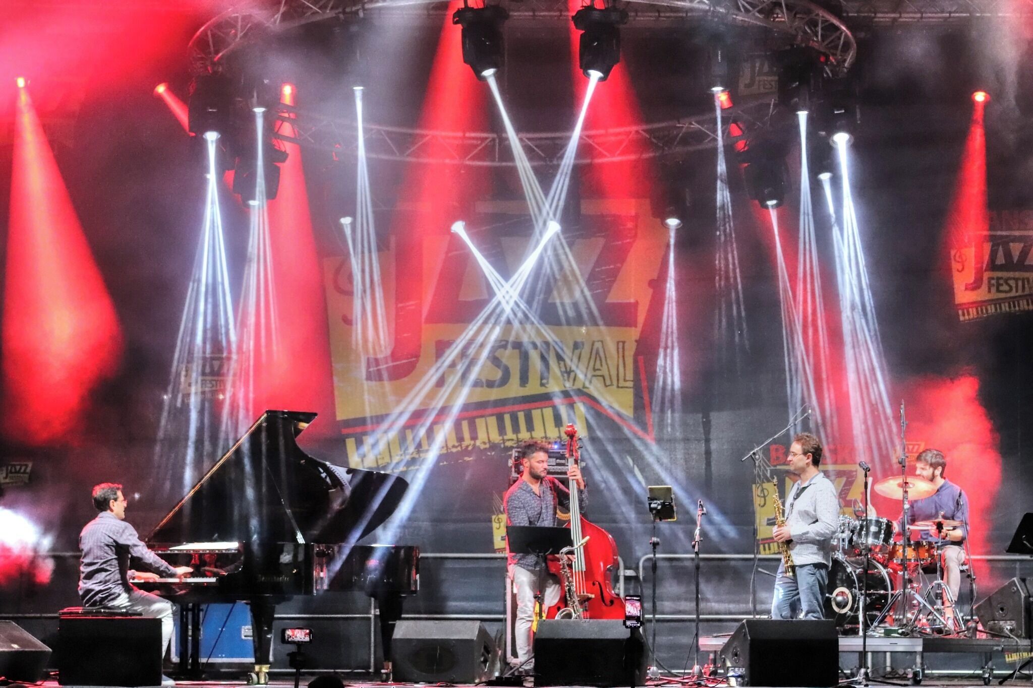 A stage with a grand piano, a double bass, a saxophone, and a drum set, indicating a live music performance.