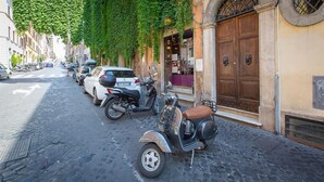 A cobblestone street lined with parked cars and scooters, a building with a large wooden door, and a tree with green leaves above the street.