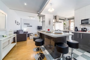 A modern kitchen with a central island, bar stools, a dining area with a sofa, and a TV.