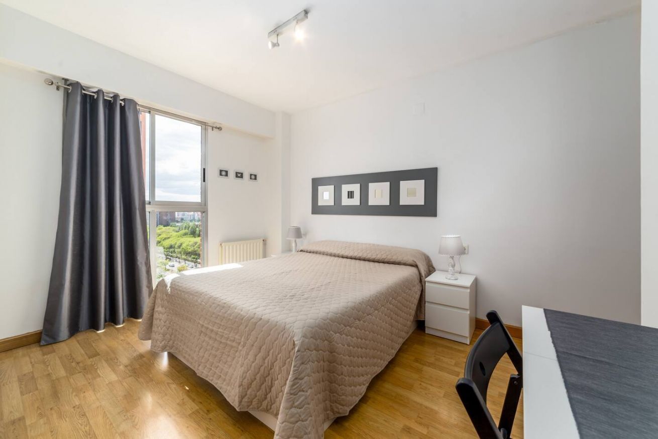 Une chambre à coucher comprenant un lit, une table de chevet, une chaise, une fenêtre avec des rideaux et une vue sur la verdure à l’extérieur.