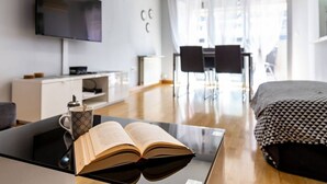 Ein Hotelzimmer mit einem Bett, einem an der Wand befestigten Fernseher, einem Schreibtisch mit Kaffeetasse und Buch sowie einem Essbereich mit Stühlen und Tisch.