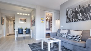 Un salon moderne avec un canapé gris, une table basse blanche et un tapis à motifs noir et blanc. On y trouve également un coin repas avec deux chaises et une table, ainsi qu'un couloir menant à une autre pièce.