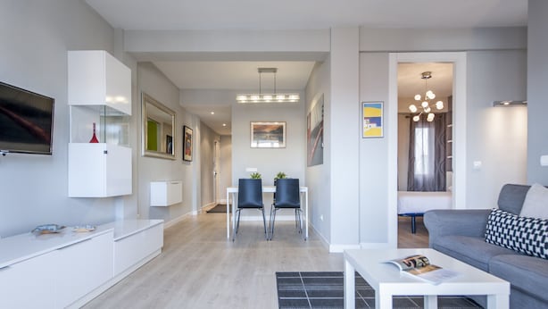 Un salon moderne avec un canapé gris, une table basse blanche et un coin repas composé de deux chaises et d’une table blanche.