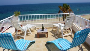 A balcony with white chairs, a table with a bottle and glasses, and a view of the beach and ocean.
