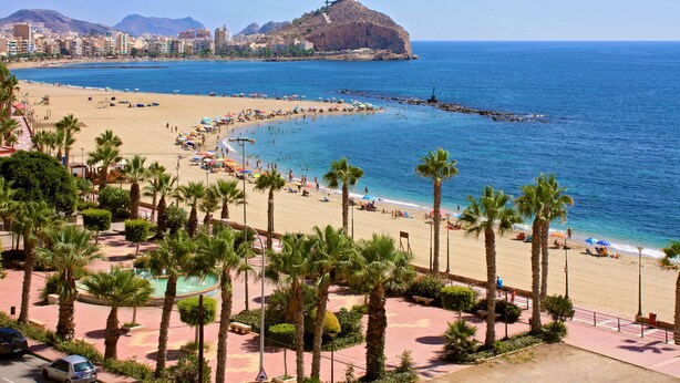 A coastal cityscape with a beach, palm trees, and a prominent hill with a fort.