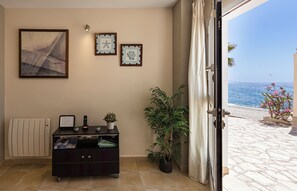 A room with a radiator, a desk with a telephone and a small plant, a framed picture on the wall, and a view of the sea through an open door.
