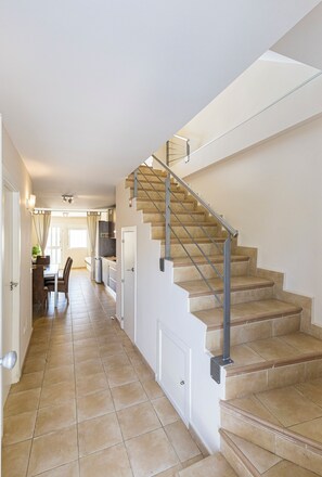 A staircase with a metal handrail leading to a landing, a dining area with chairs and a table, and a kitchen with cabinets and a refrigerator.