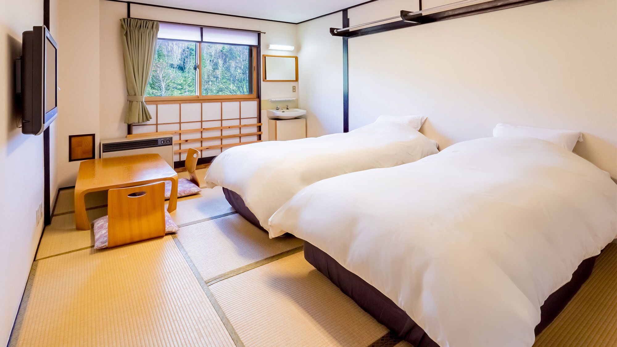 A hotel room with two beds, a small table, a TV, a window with a view of trees, and a bathroom with a sink and mirror.