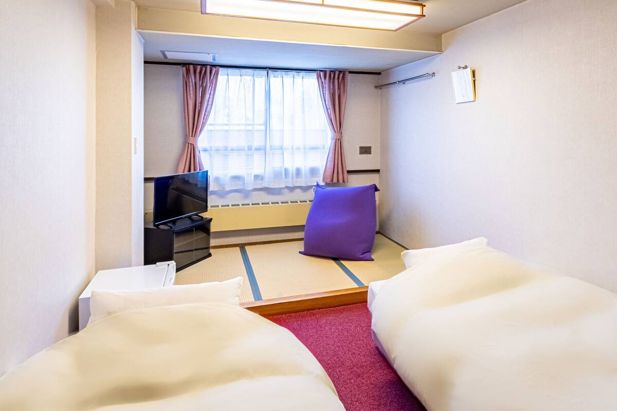 Hotellihuone, jossa on sänky, pieni pöytä, tuoli, televisio ja ikkuna, jossa on verhot.