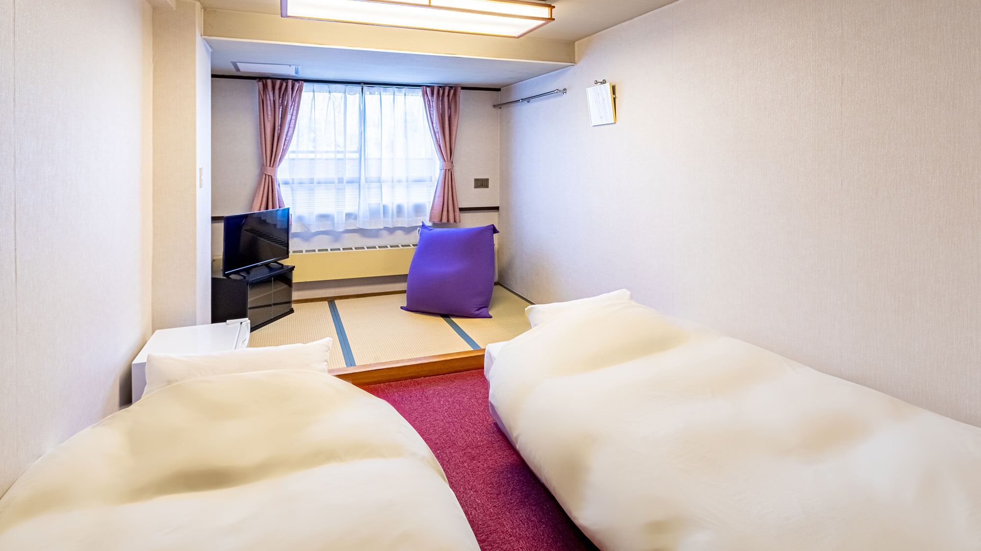 A hotel room with a bed, a television, a purple cushion, and a window with curtains.