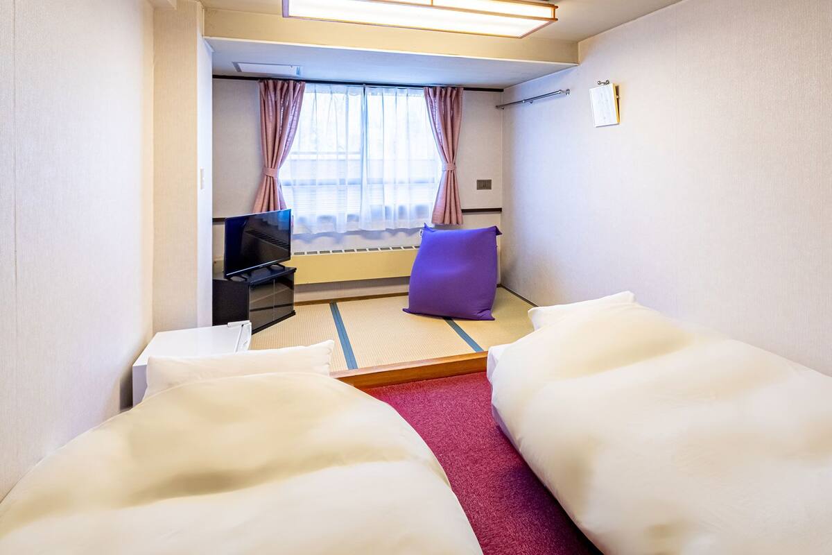 Hotellihuone, jossa on sänky, televisio, violetti koristetyyny ja ikkuna, jossa on verhot.