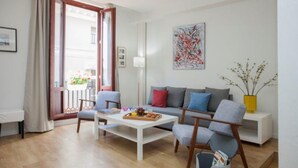 A modern living room with a sofa, two armchairs, a coffee table, and a small side table. There is a window with curtains, a painting on the wall, and a vase with branches on a side table.