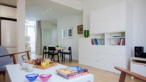 A modern living room with a dining area, a bookshelf, and a kitchen visible in the background.