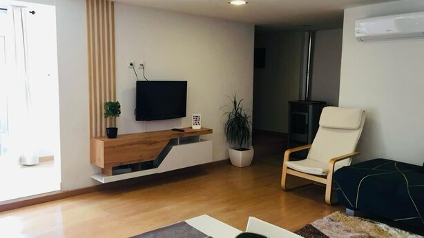 A modern living room with a flat-screen TV mounted on a wooden console, a comfortable chair, a potted plant, and a large window providing natural light.