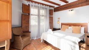 A bedroom with a large bed, a wicker chair, wooden cabinets, and a window with sheer curtains.