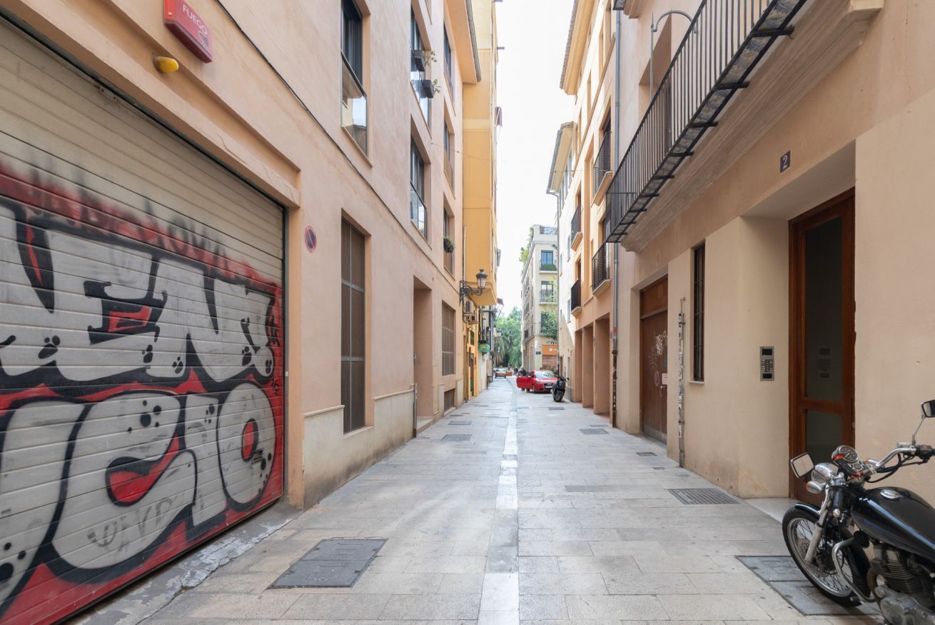 A narrow alleyway with a closed garage door on the left, a motorcycle parked on the right, and buildings with balconies and windows.